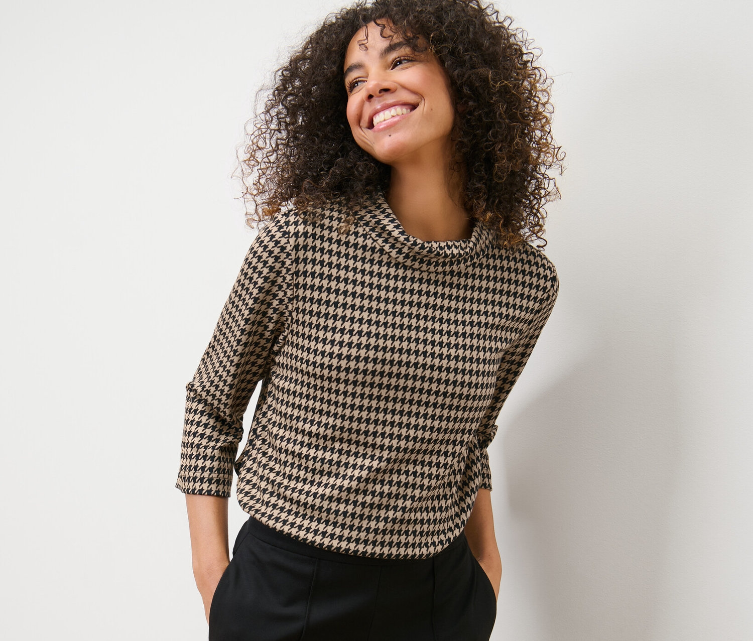 Žena s kudrnatými vlasy pózuje v mikině s 3/4 rukávem a kohoutí stopou a černých kalhotách.