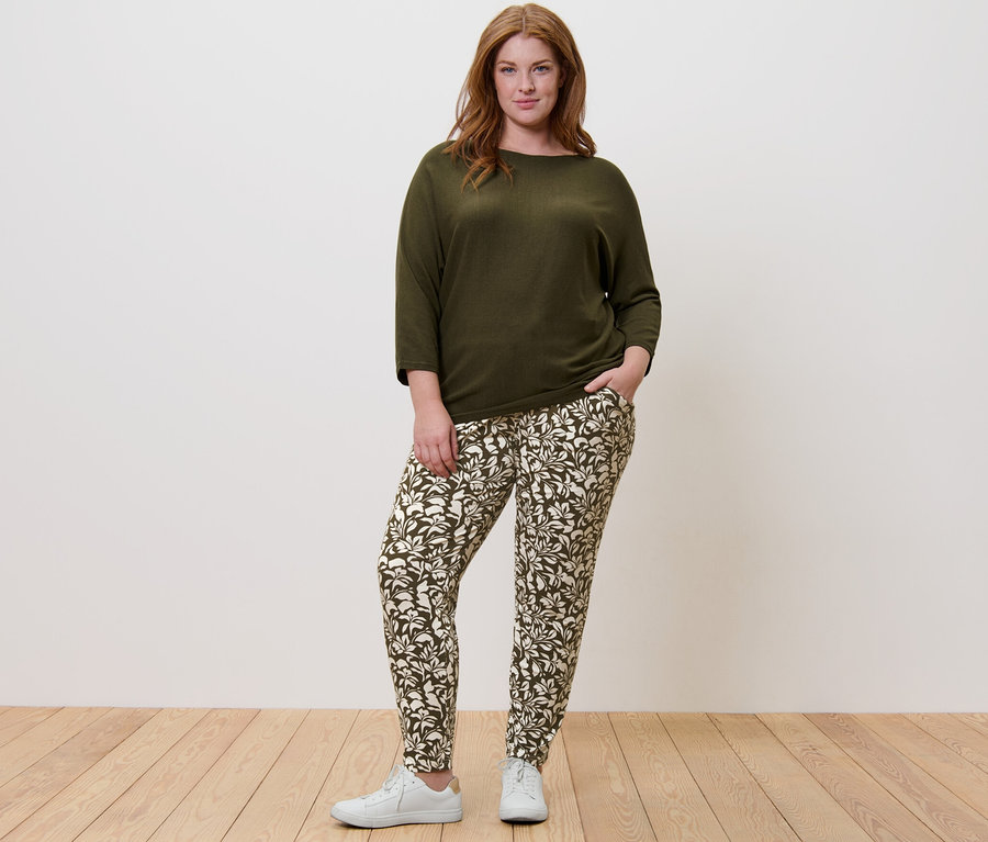 Žena stojí a má na sobě zelený svetr z jemného úpletu a vzorované žerzejové kalhoty.