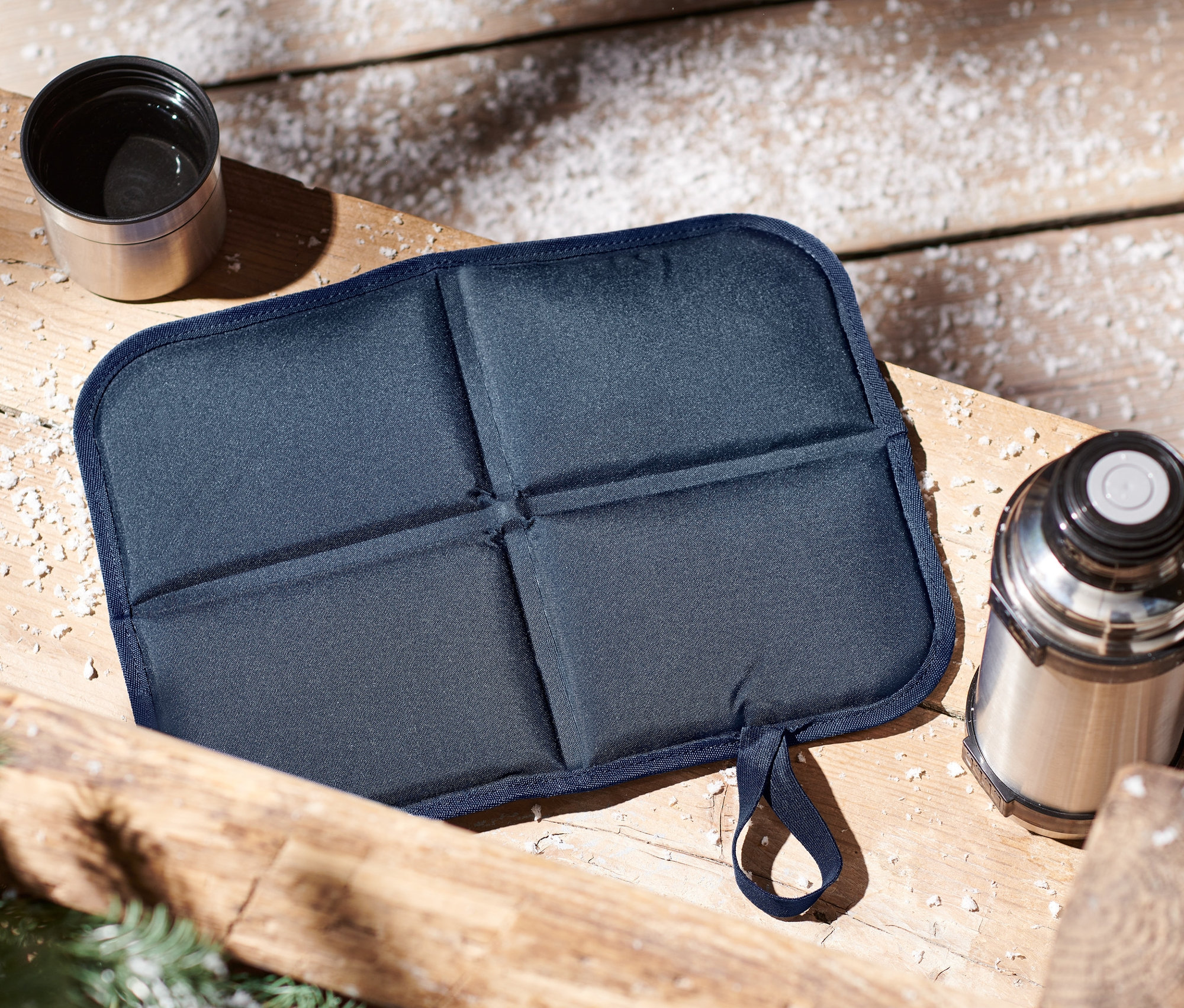 Skládací termopolštářek na sezení a termoska na dřevěném stole.