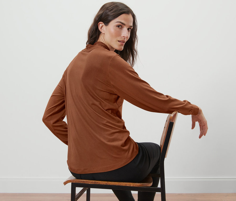 Žena sedí na židli a má na sobě koňakově hnědé tričko s dlouhým rukávem a výstřihem do V.