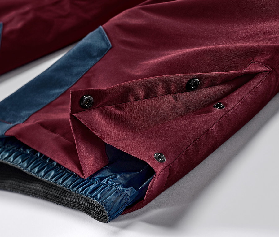 Detail vínově červených lyžařských a snowboardových kalhot s modrými akcenty a zapínáním na knoflíky.