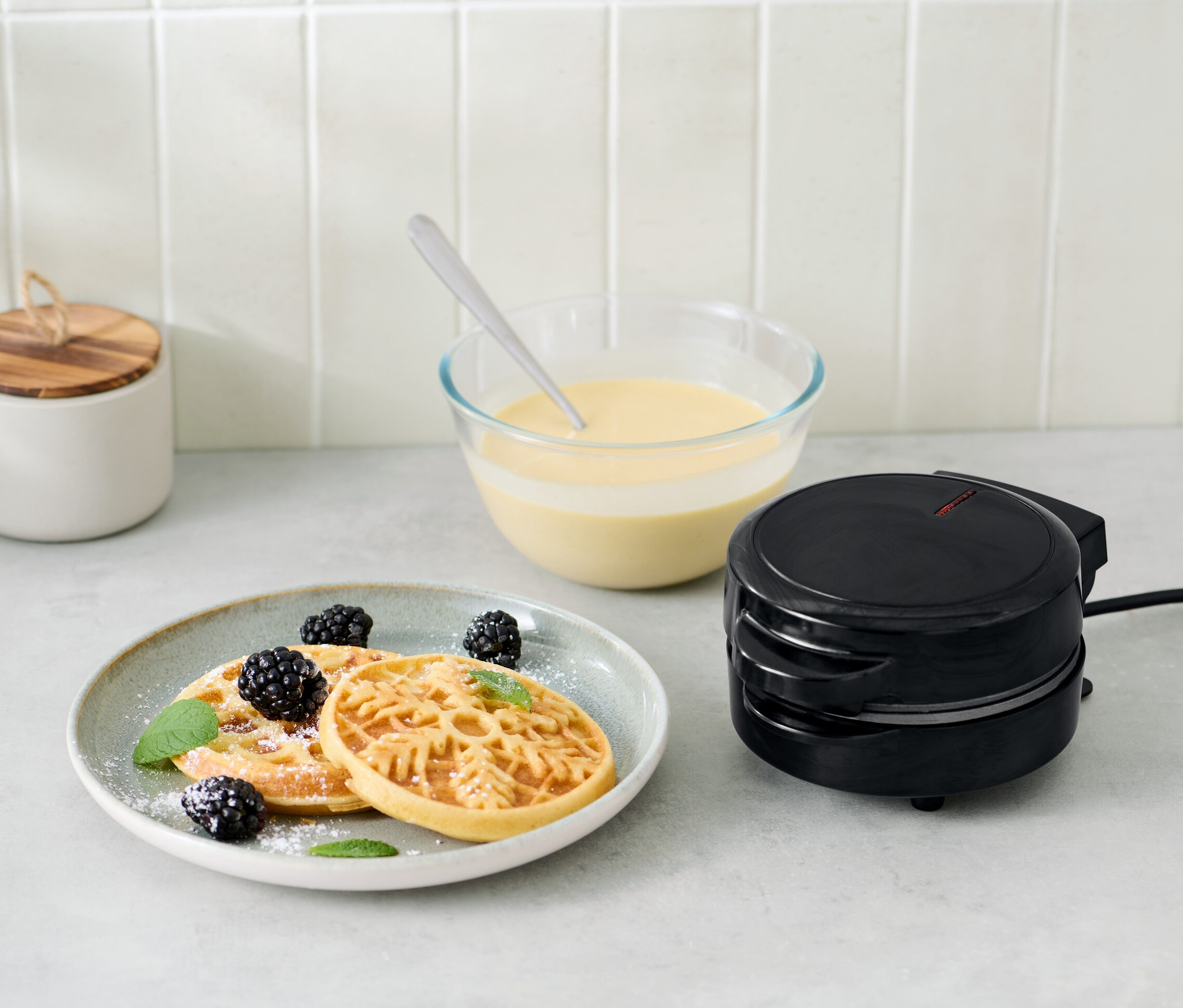 Mini vaflovač vedle talíře s vaflemi, ostružinami a lístky máty.