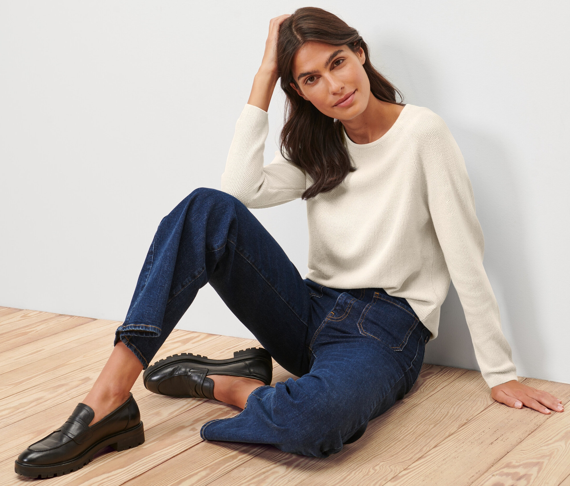 Žena sedí na dřevěné podlaze a má na sobě krémově bílý pletený svetr.