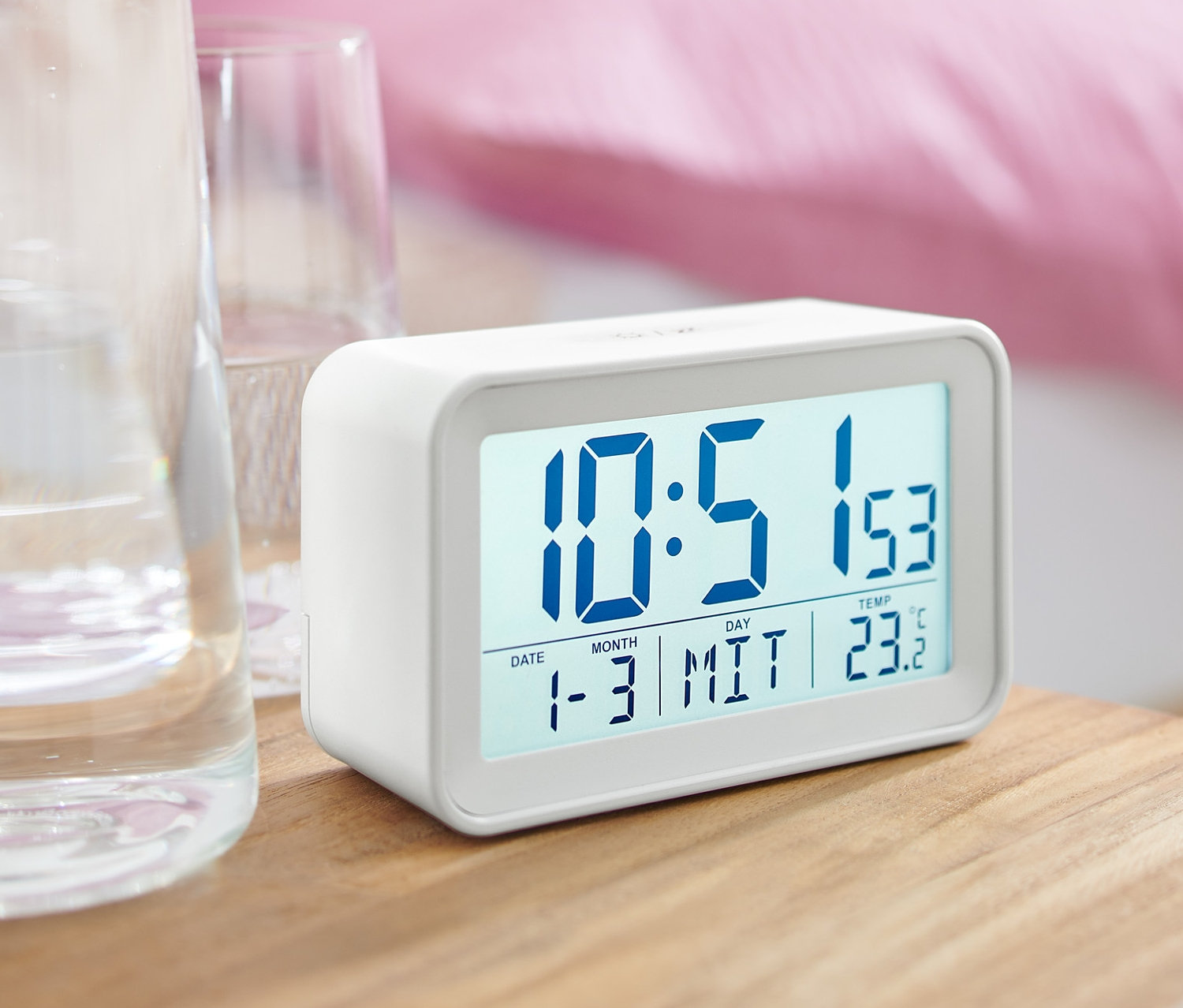 Bílý digitální LCD budík na nočním stolku vedle sklenice s vodou.