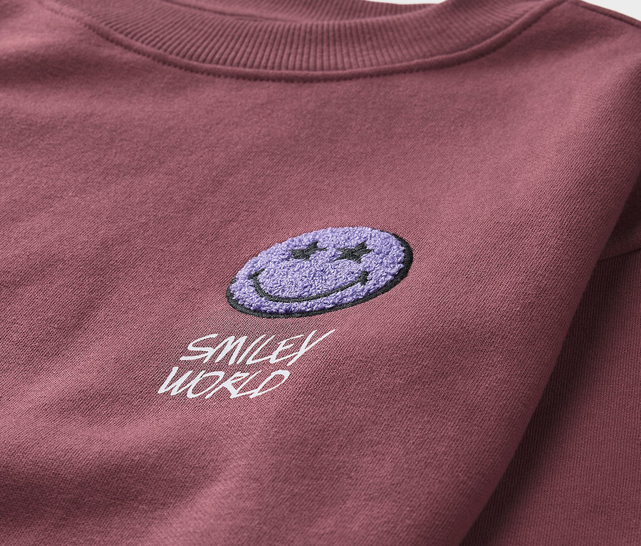 Detail bordó dětské mikiny SMILEYWORLD® s nášivkou fialového smajlíka.