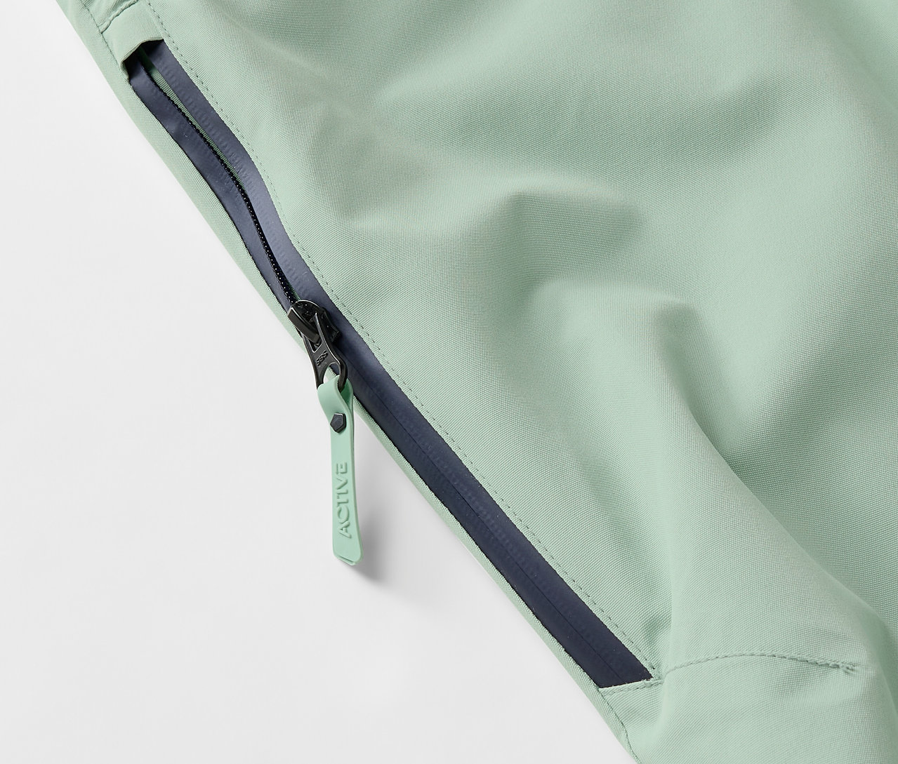 Detail světle zelené bundy se zipem a tmavě modrým lemem.