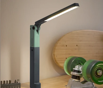 Na dřevěném stole stojí LED pracovní lampa, šroubovák, sada bitů a skateboardové příslušenství.