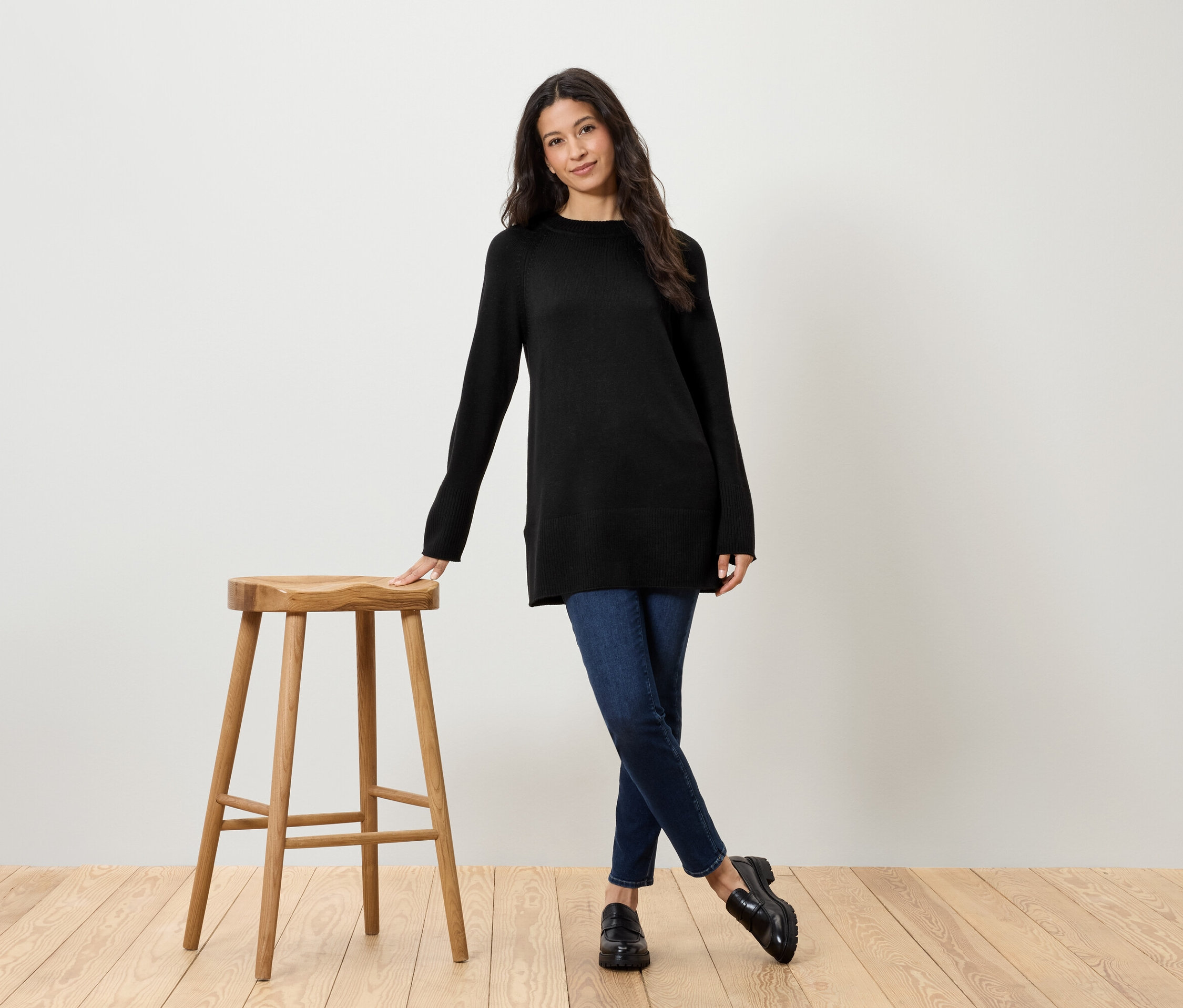 Žena v černém svetru a džínách stojí vedle dřevěné stoličky.