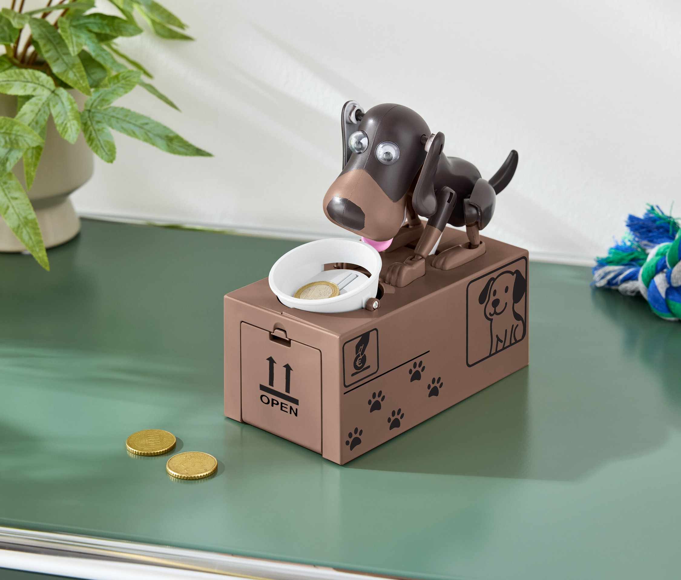 Hnědá kasička ve tvaru psa s mincí v misce a dvěma mincemi na zeleném stole.