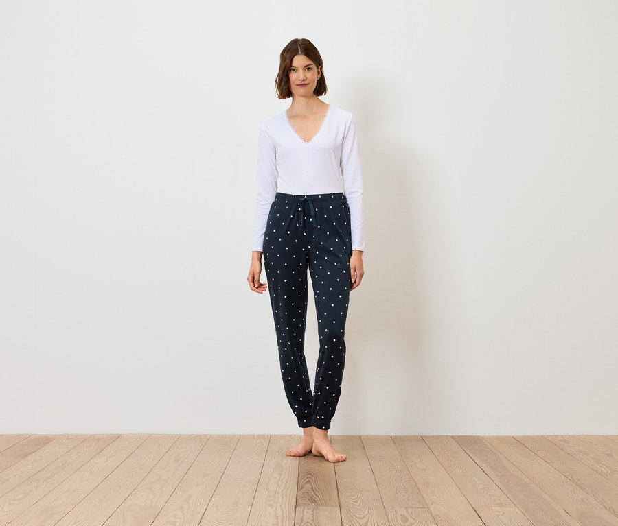 Žena stojí a má na sobě bílé tričko s dlouhým rukávem a tmavě modré kalhoty se vzorem.