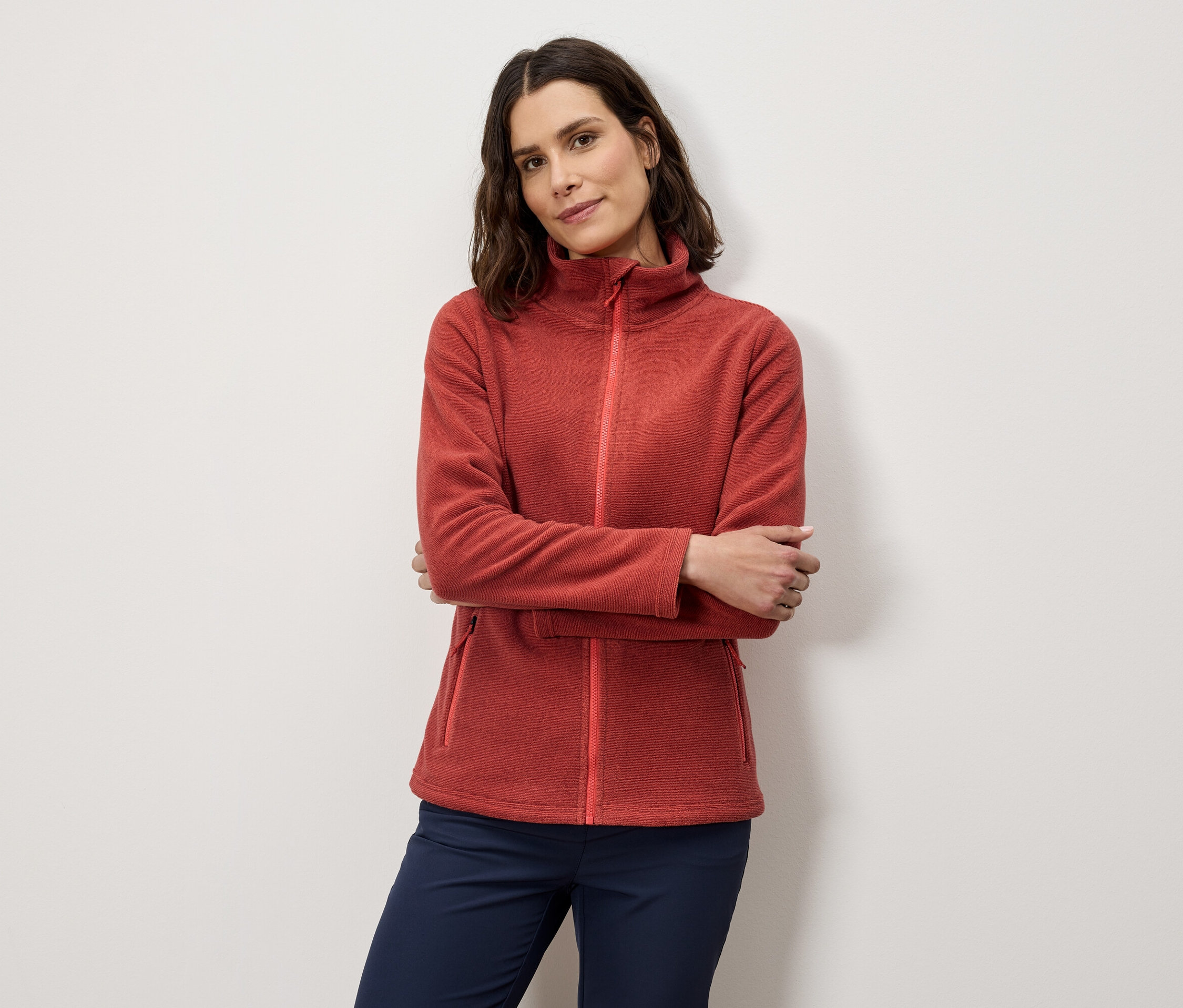 Žena se zkříženýma rukama pózuje v oranžové fleecové bundě a tmavě modrých kalhotách.