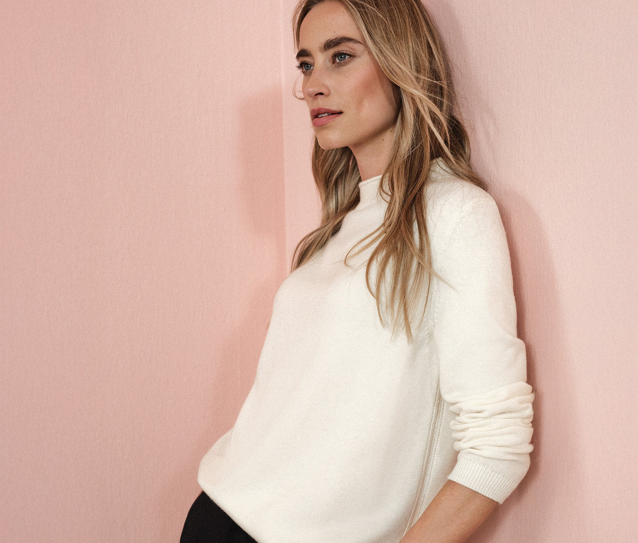 Žena se opírá o růžovou stěnu a má na sobě béžový pletený svetr s rolovaným lemem Street One.