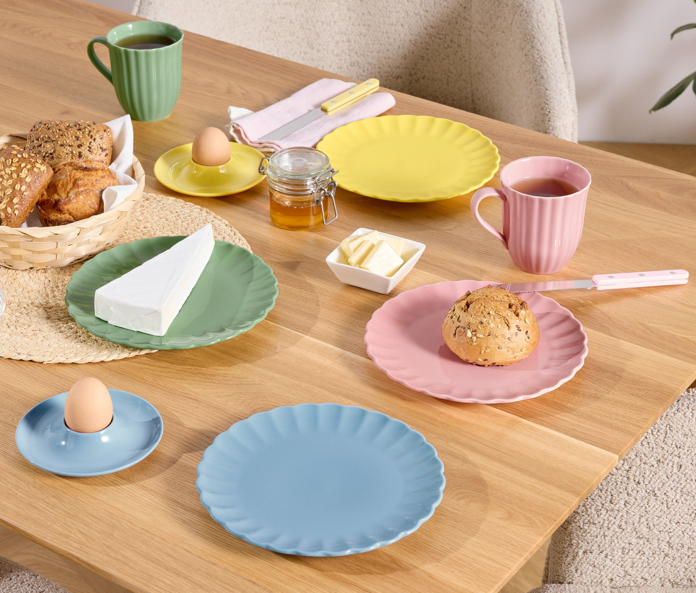 Na dřevěném stole jsou prostřeny barevné talíře, šálky a misky s jídlem, včetně chleba v košíku, vajíčka v kalíšku a kousku sýra.