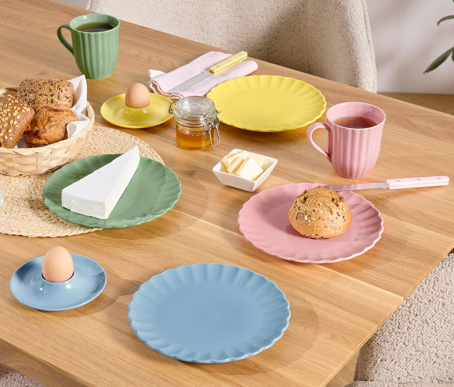 Na dřevěném stole jsou prostřeny barevné talíře, šálky a misky s jídlem, včetně chleba v košíku, vajíčka v kalíšku a kousku sýra.