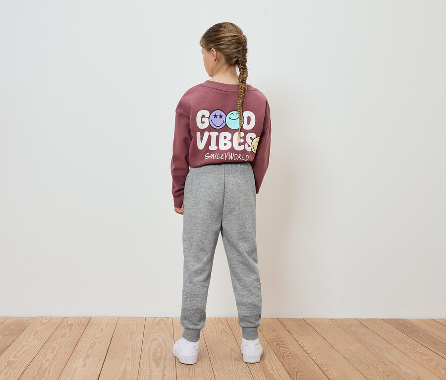Dívka v bordó mikině s nápisem Good Vibes a šedých teplácích stojí zády k fotoaparátu.