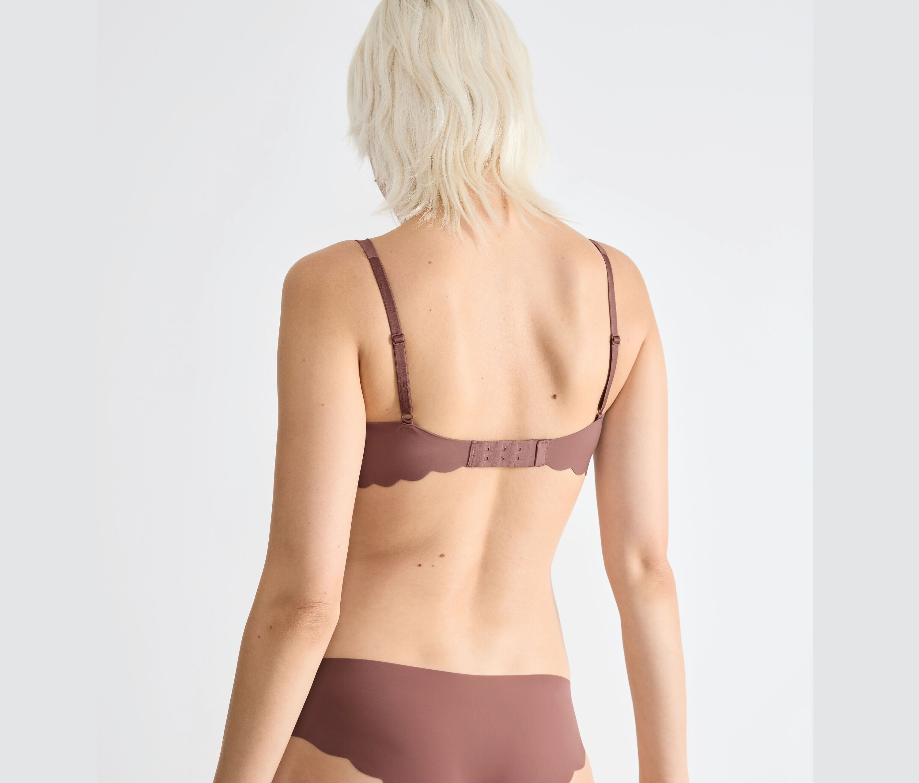 Pohled zezadu na ženu s blond vlasy v hnědé podprsence sloggi ZERO Microfibre 2.0 Soft Bra a kalhotkách.