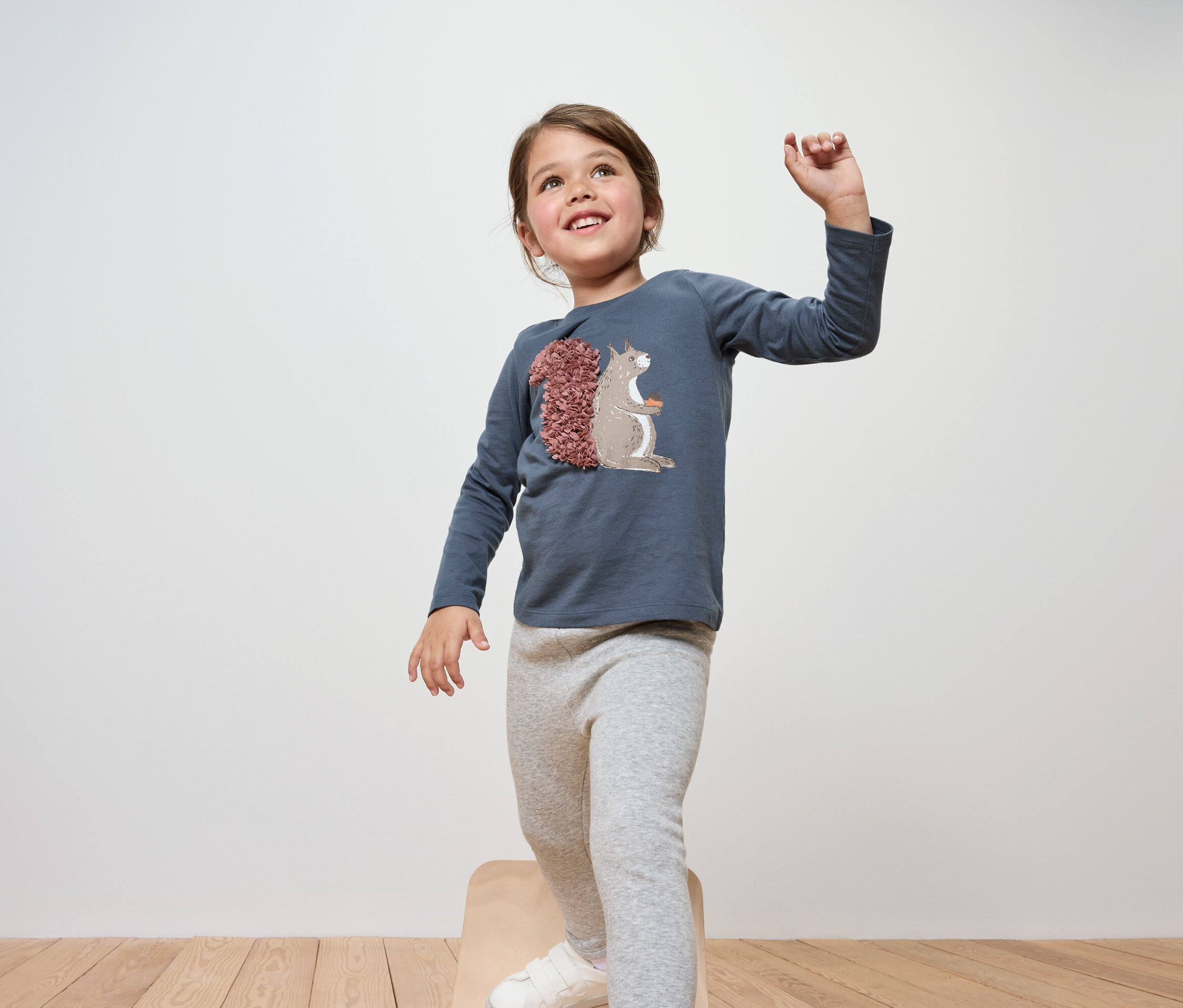 Usmívající se dívka stojí na dřevěné stoličce, má na sobě modré tričko s dlouhým rukávem a šedé legíny.