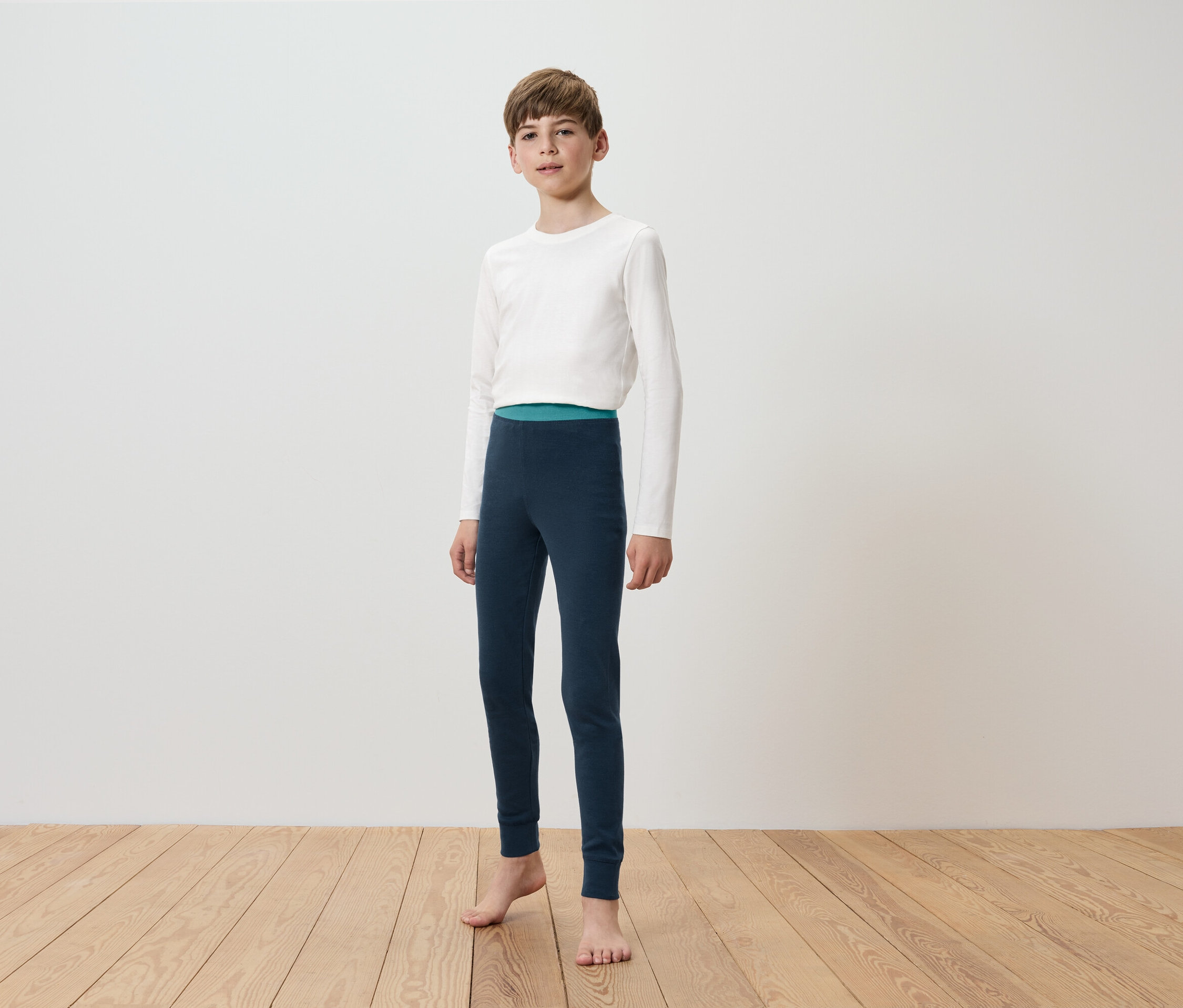 Chlapec v bílém tričku s dlouhým rukávem a tmavě modrých legínách stojí na dřevěné podlaze před bílou stěnou.