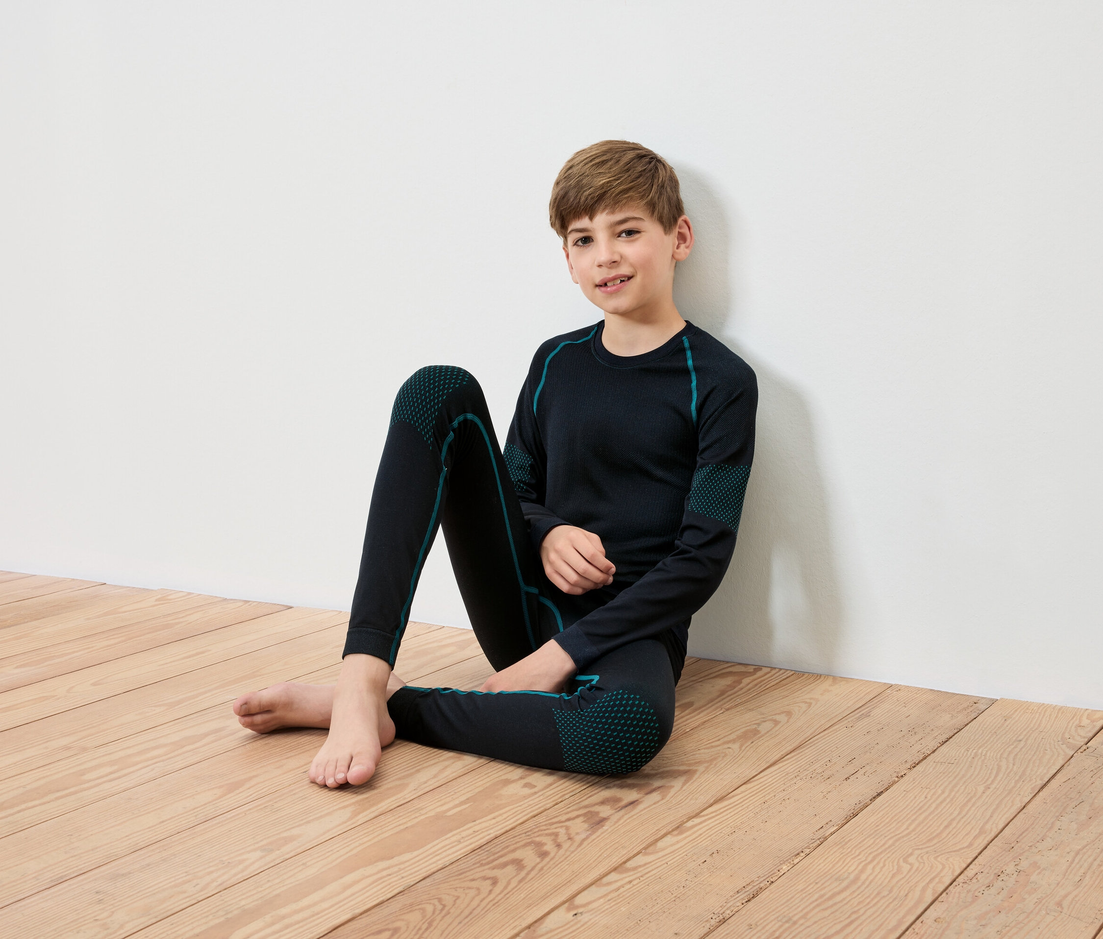 Chlapec sedí na dřevěné podlaze opřený o bílou zeď, má na sobě černé termoprádlo s tyrkysovými detaily.