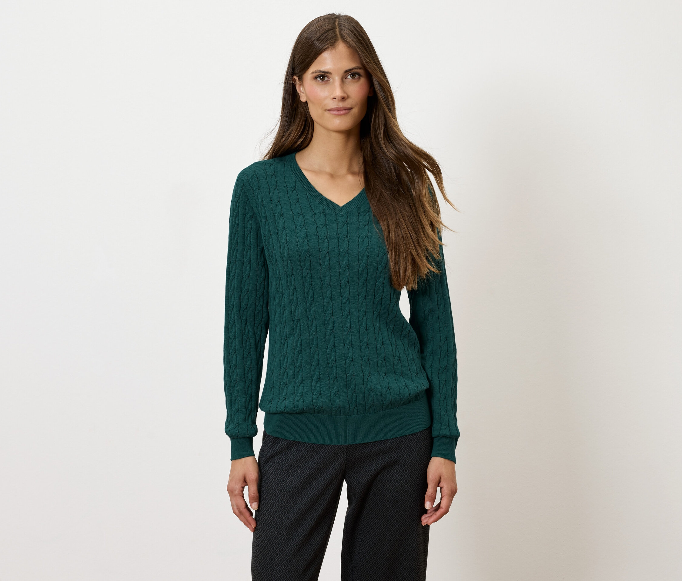 Žena s dlouhými hnědými vlasy má na sobě zelený svetr s véčkovým výstřihem a černé kalhoty se vzorem.