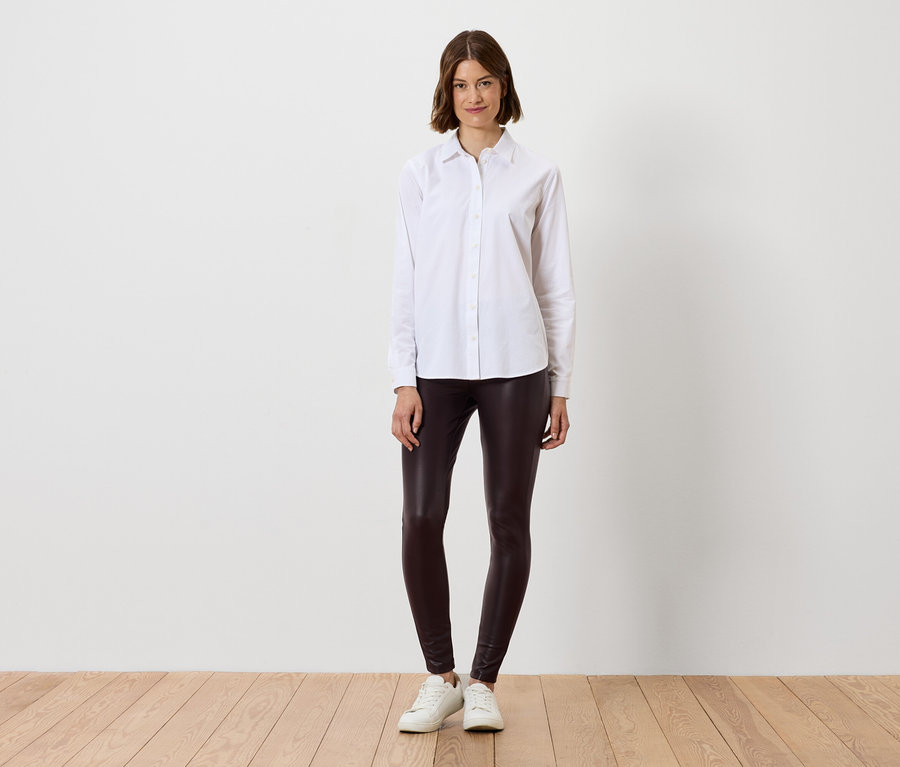 Žena v bílé košili a hnědých legínách stojí na dřevěné podlaze.