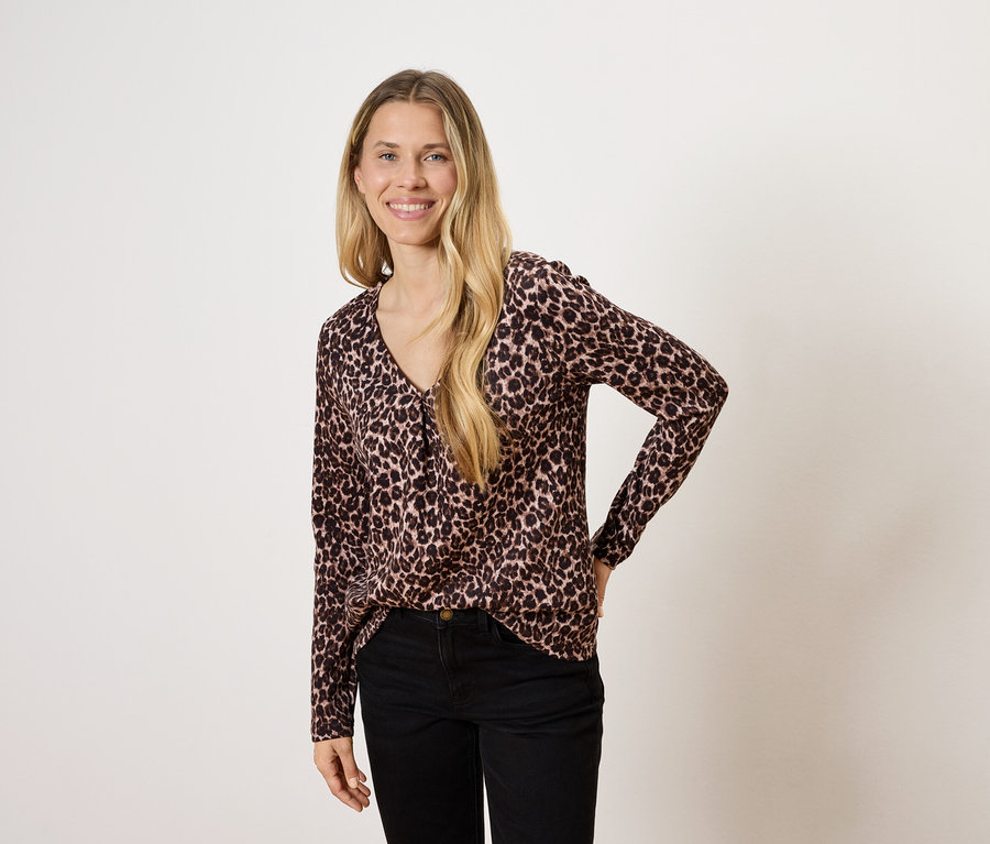 Žena s dlouhými blond vlasy pózuje v halence se zvířecím vzorem a černých kalhotách.