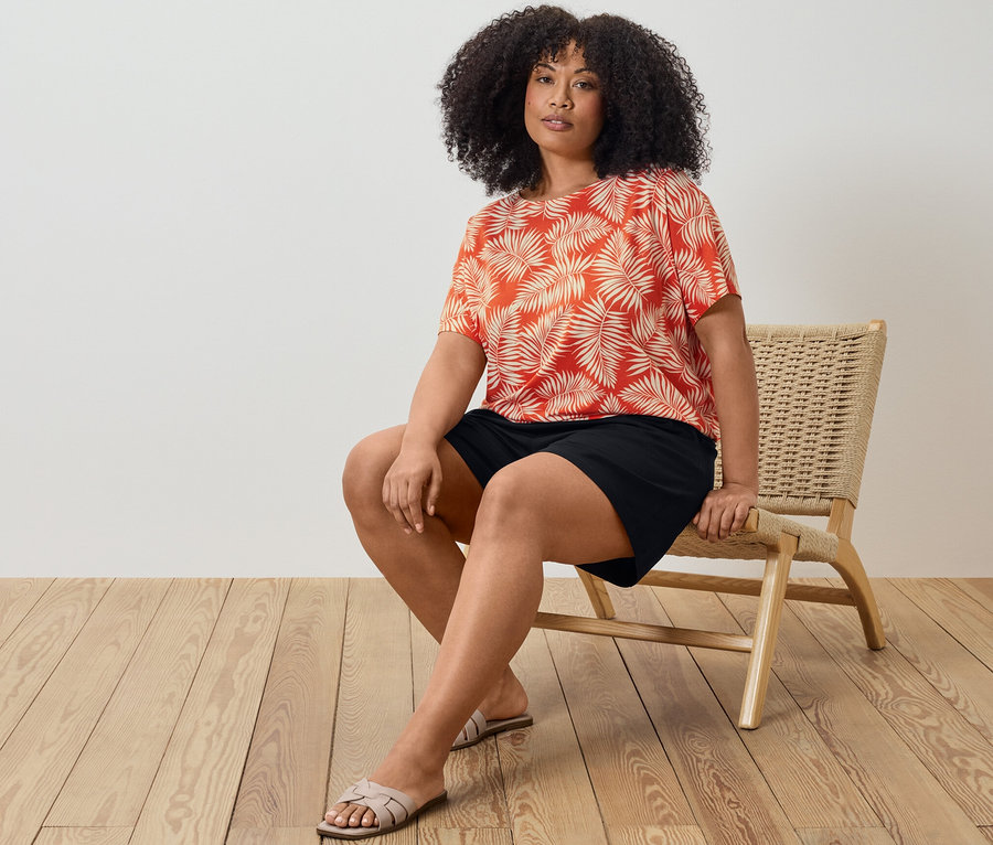 Žena sedí na židli a má na sobě halenkové triko s potiskem a černé žerzejové šortky.