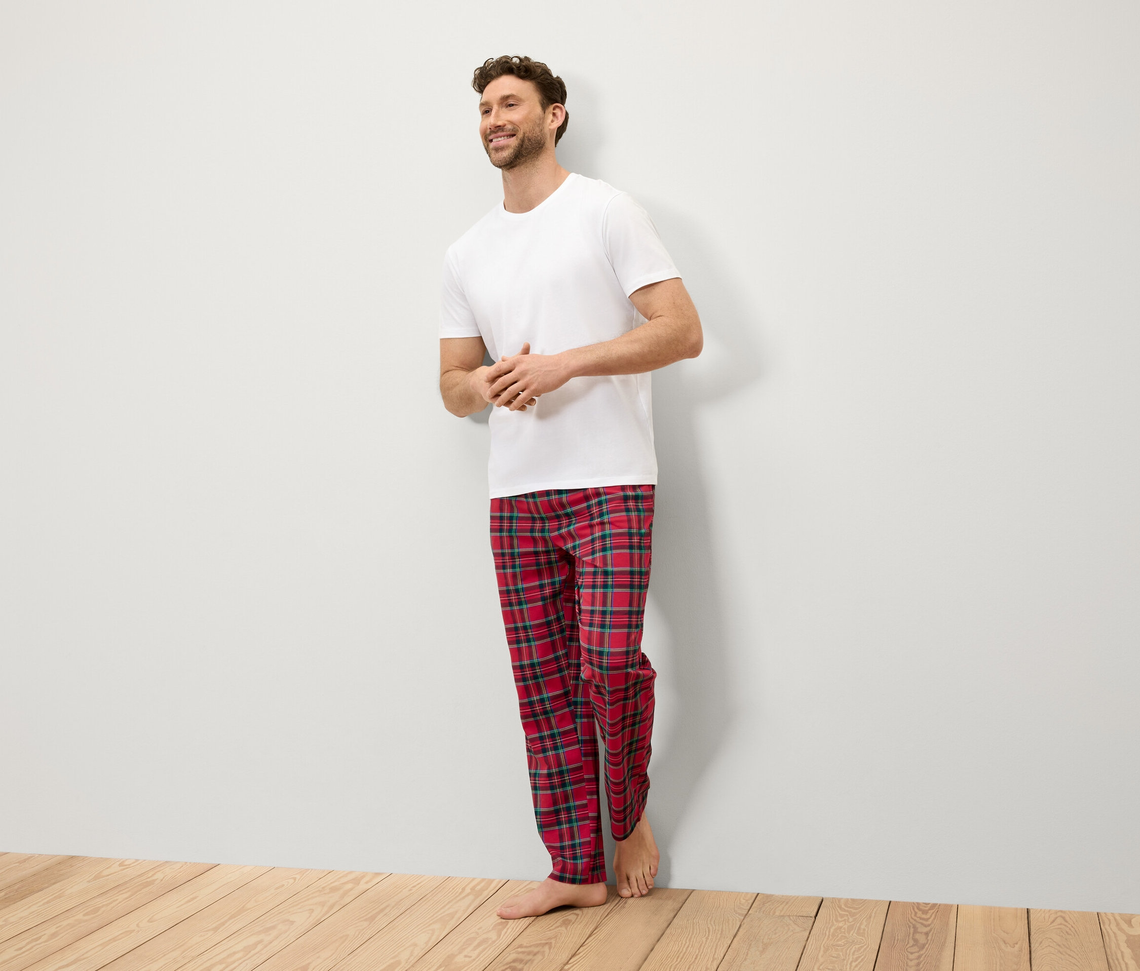 Usmívající se muž stojí u stěny v bílém tričku a červených kostkovaných kalhotách.