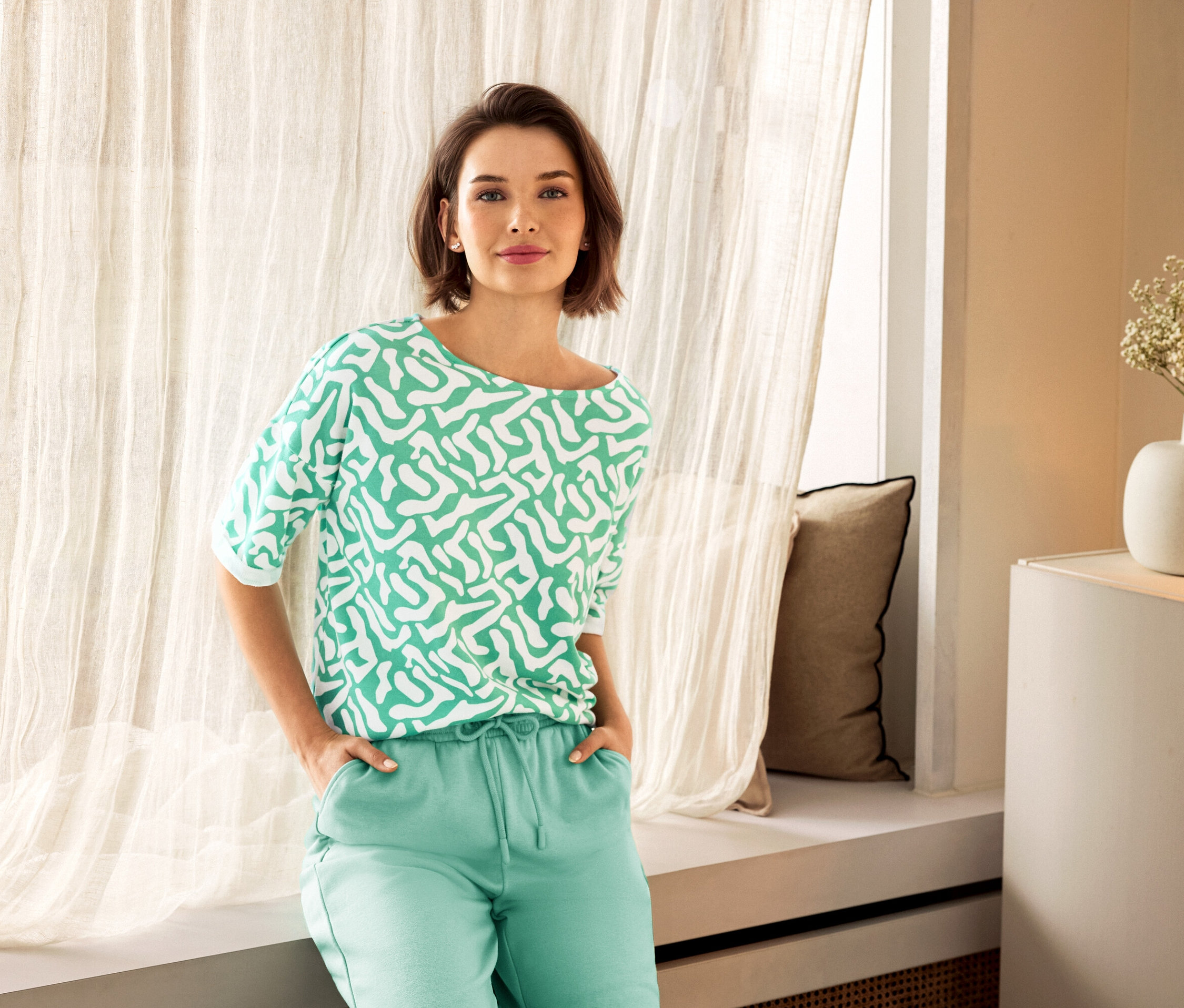 Žena v zelených kalhotách a vzorované halence pózuje u okna. V pozadí je vidět váza.