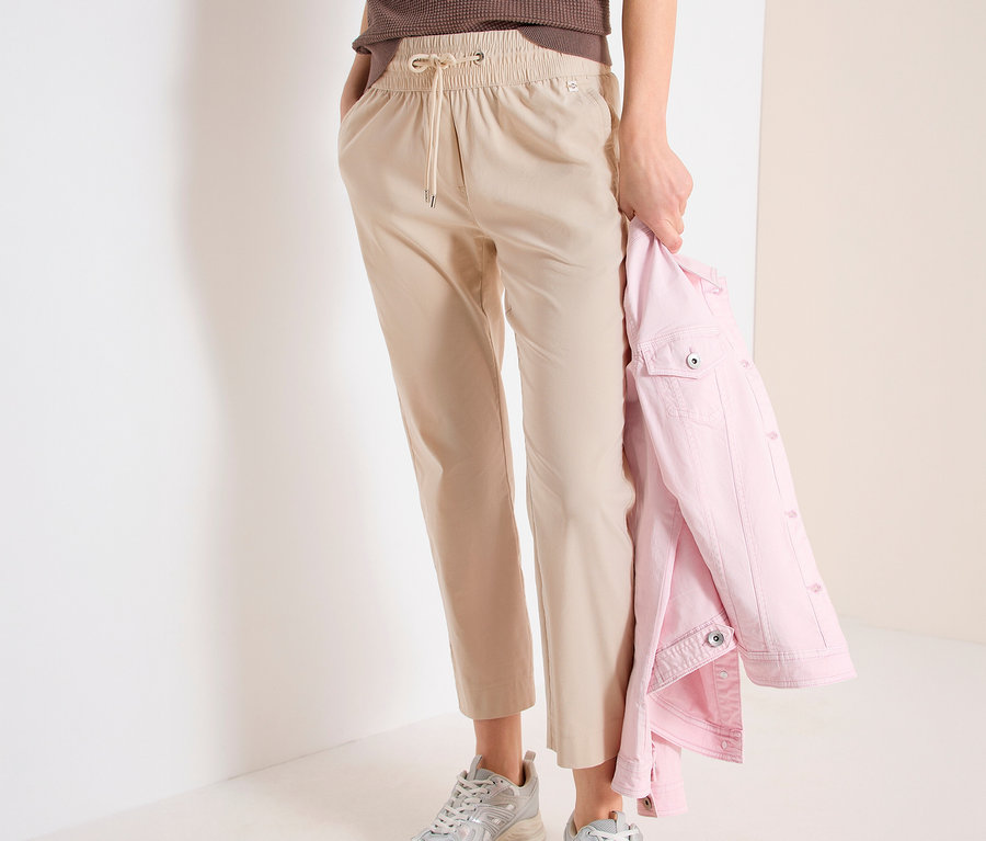 Žena v kalhotách joggpant CECIL »Tracey«, hnědém topu a stříbrných teniskách drží růžovou bundu.