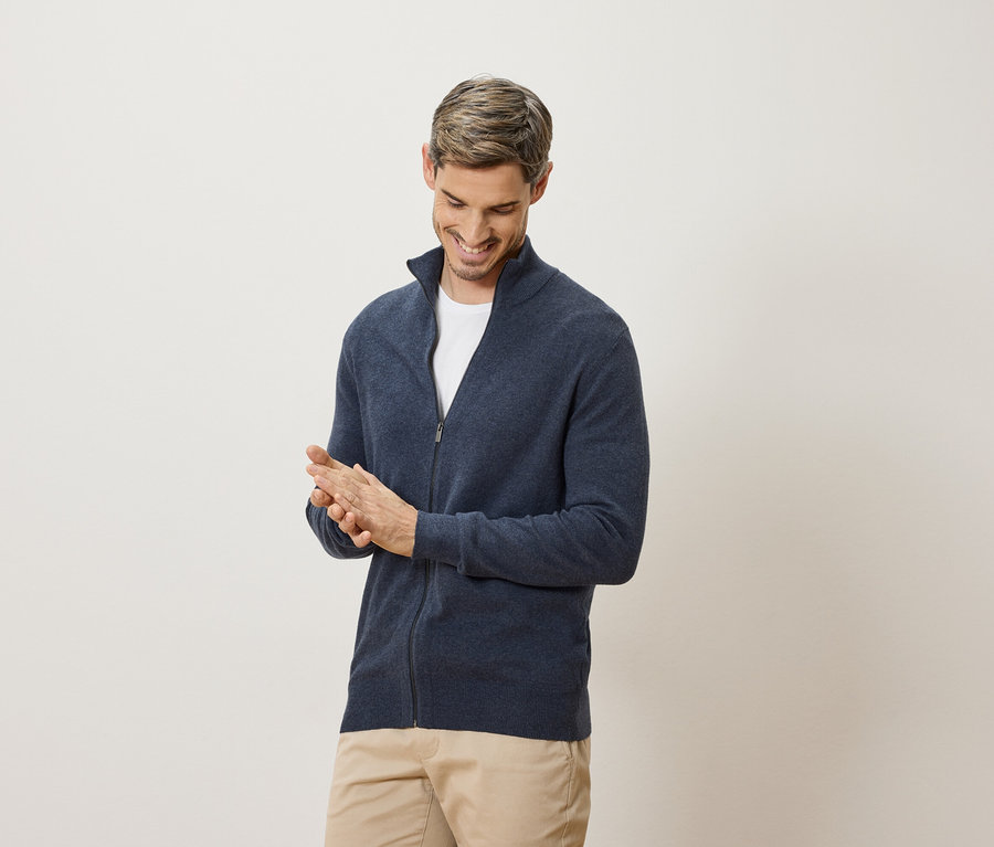 Muž se usmívá a tře si ruce. Má na sobě tmavě modrý kardigan a béžové kalhoty.