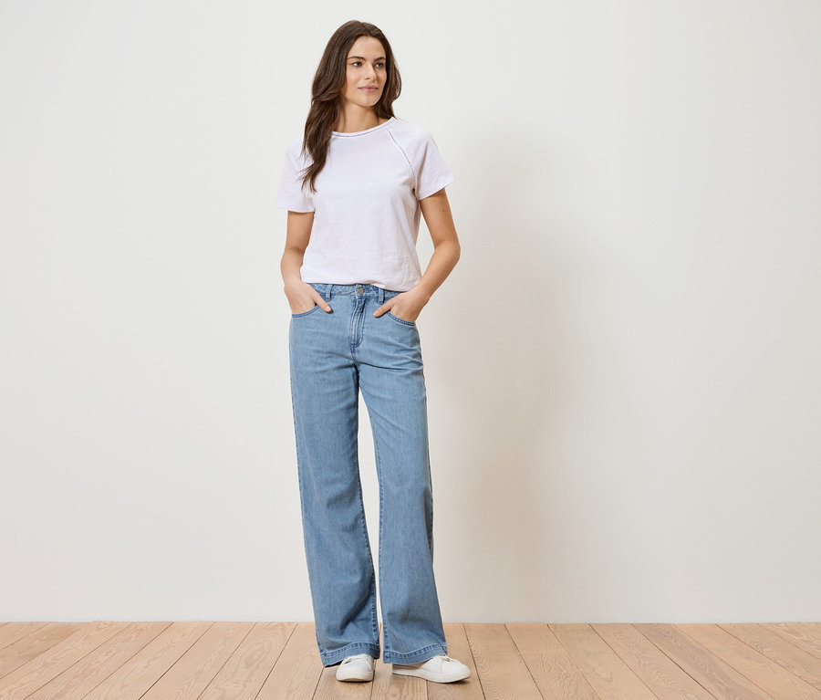 Žena v bílém tričku a modrých džínách stojí před bílou zdí.