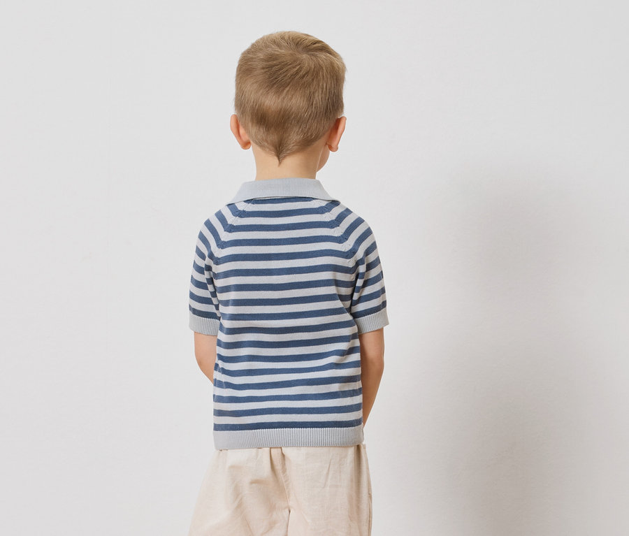 Pohled zezadu na dítě s blond vlasy v modro-bíle pruhovaném polo tričku a béžových šortkách na bílém pozadí.