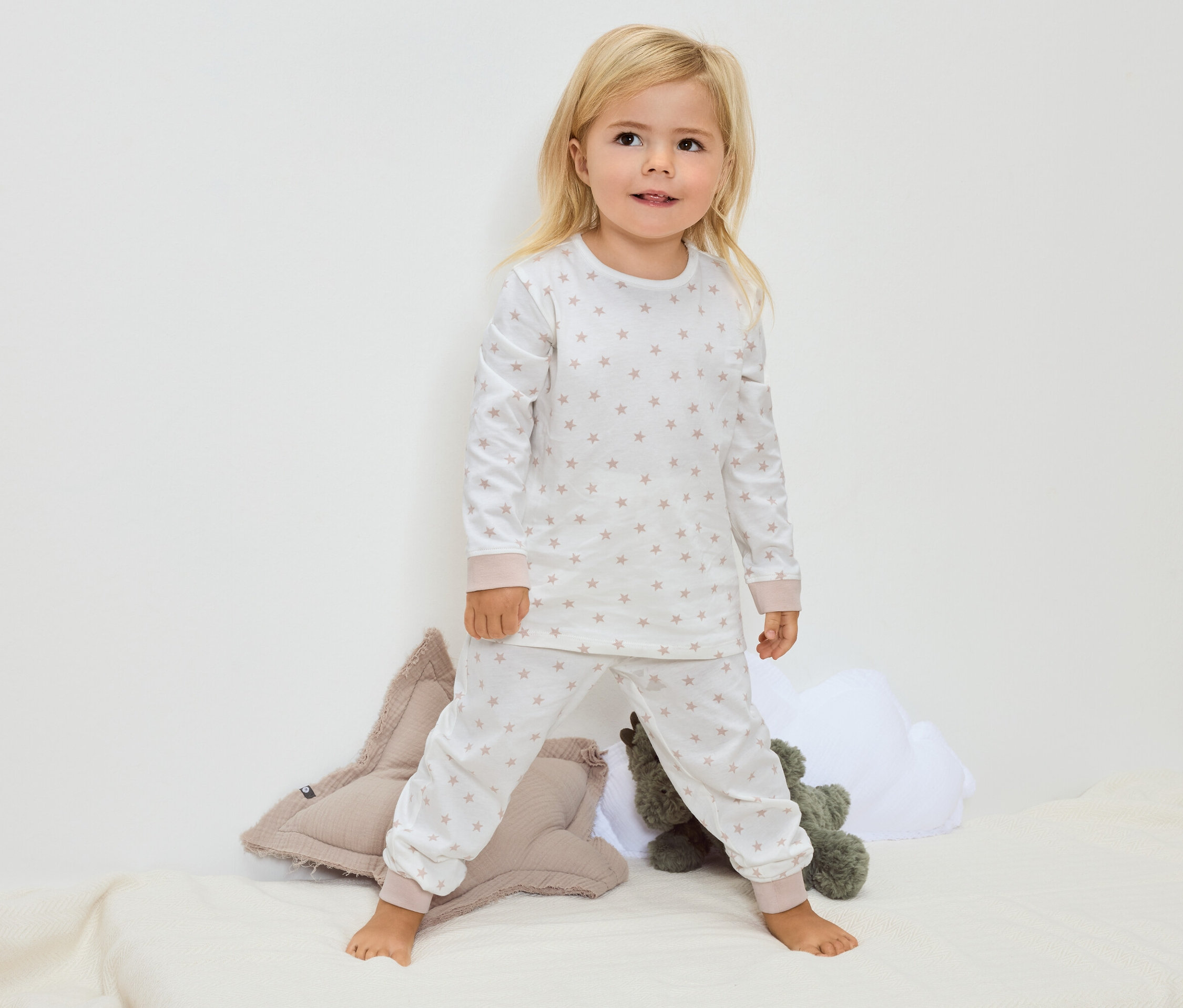 Malá blondýna stojí bosá na dece a má na sobě bílé pyžamo s hvězdičkami.