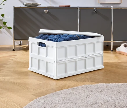 Na dřevěné podlaze stojí bílý skládací box s modrým obsahem, za ním šedá skříňka a boty.