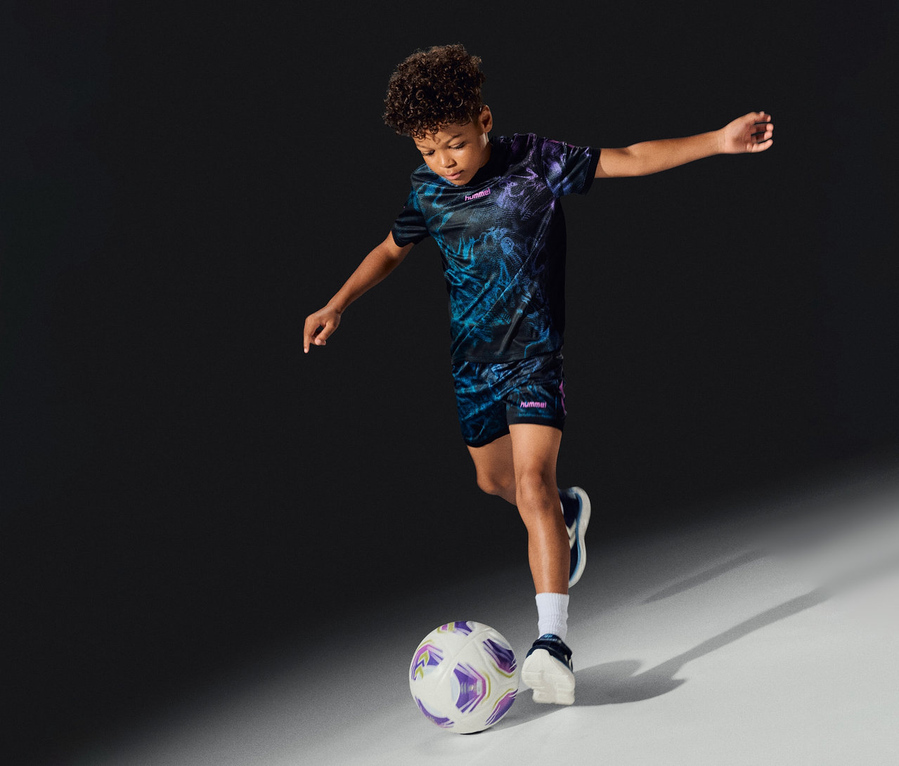 Chlapec v soupravě hummel® »HML Blaze Graphic Set Kids« hraje fotbal a kope do bílo-fialového míče.