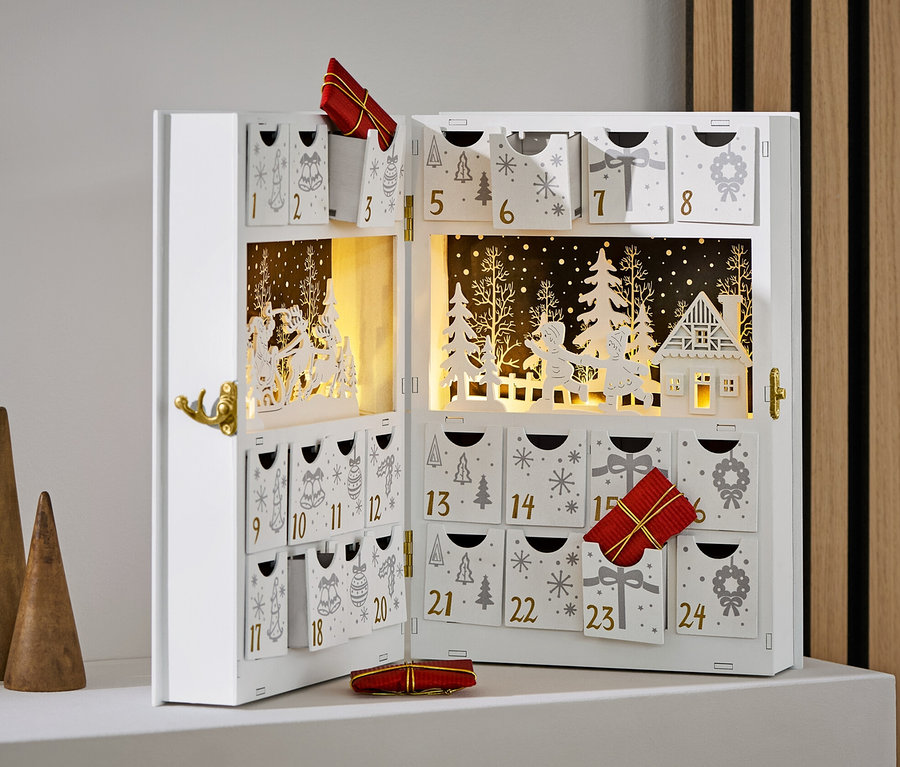 Otevřený bílý adventní kalendář s ozdobami a dárky. Stojí vedle tří dřevěných kuželů.