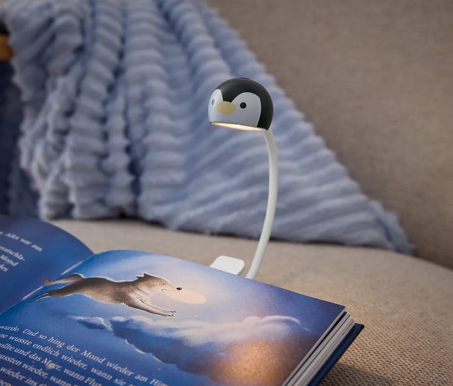 Otevřená kniha s tmavě modrým obalem leží na béžové pohovce, osvětlená klipovou lampou. Lampa má bílé flexibilní rameno a černo-bílou hlavu ve tvaru tučňáka, která vrhá světlo na stránky knihy. Pravá strana zobrazuje ilustraci vlka skákajícího přes zataženou, modrou oblohu. Světle modrá, texturovaná deka je přehozená přes opěradlo pohovky v pozadí.