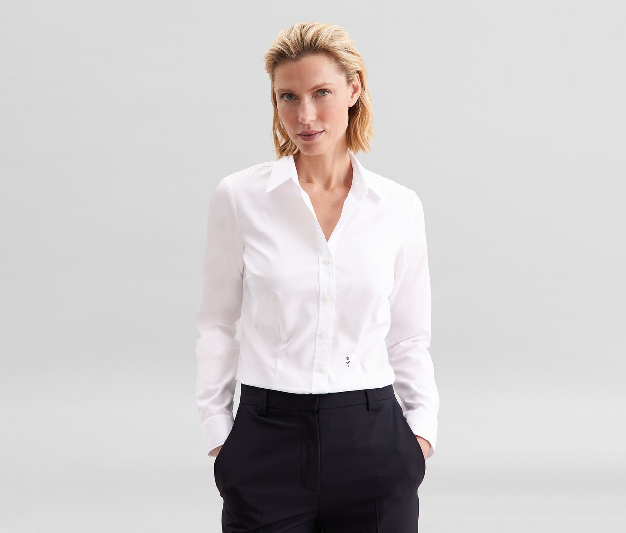 Blondýnka stojí s rukama v kapsách, oblečená v bílé halence slim fit non iron Seidensticker a tmavých kalhotách.