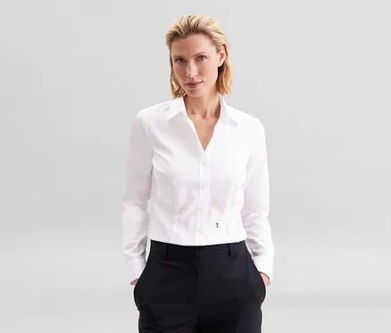 Blondýnka stojí s rukama v kapsách, oblečená v bílé halence slim fit non iron Seidensticker a tmavých kalhotách.