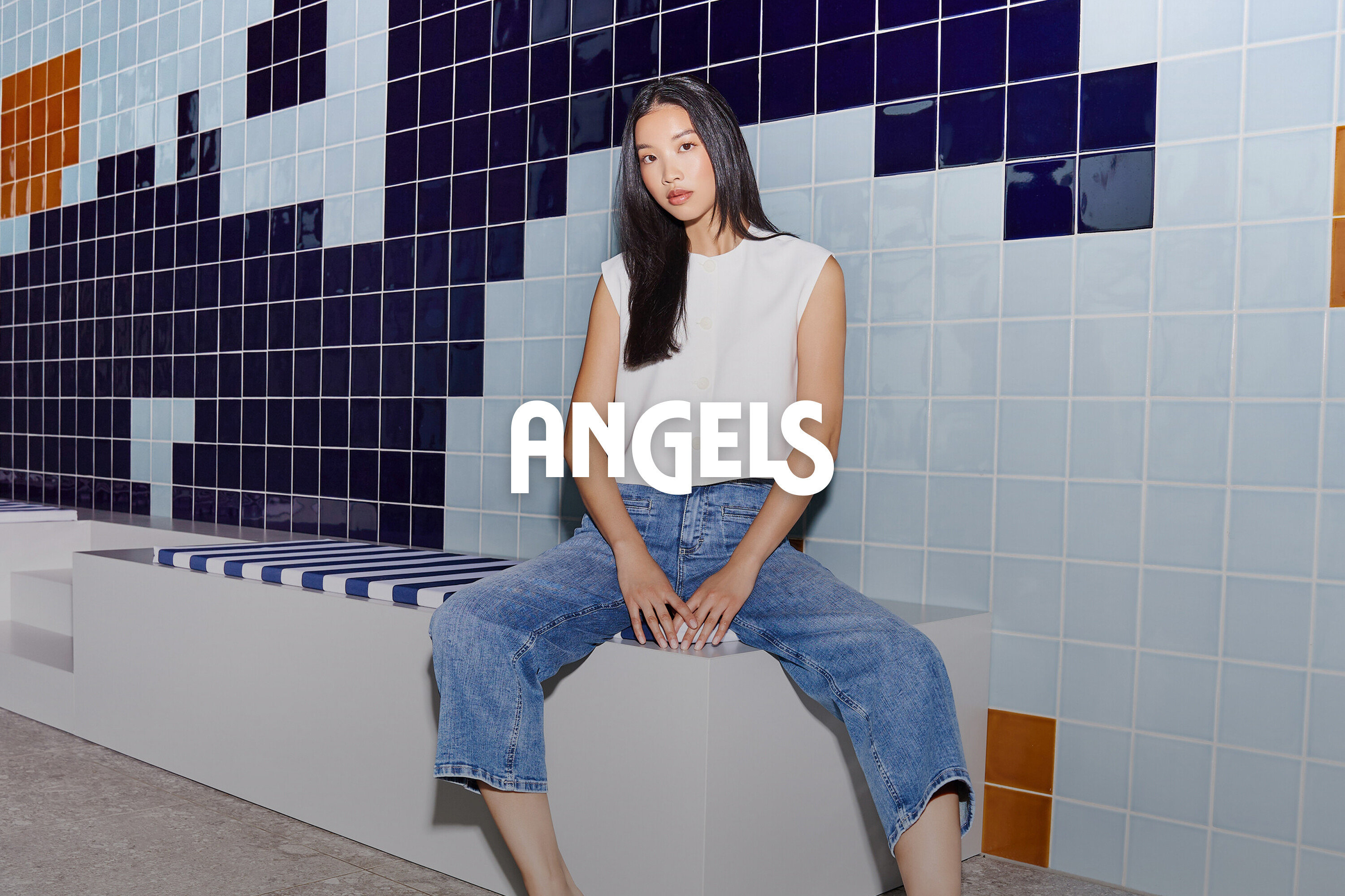 Žena sedí na lavičce před stěnou s modrými dlaždicemi. Má na sobě džíny a bílý top.