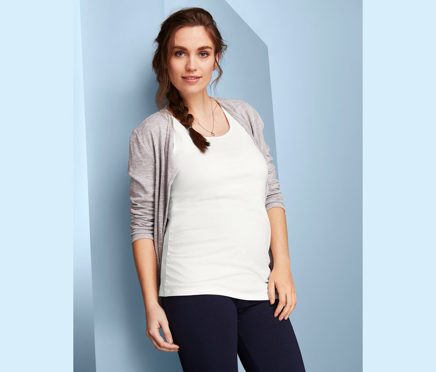 Těhotná žena pózuje v bílém topu z dvoukusového setu těhotenských topů a v šedém svetru.