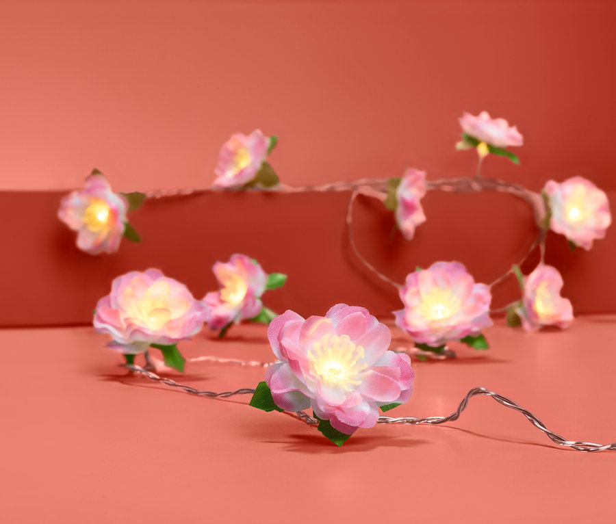Světelný řetěz s LED světly ve tvaru růžových květů s bílým středem a zelenými lístky na korálovém pozadí.