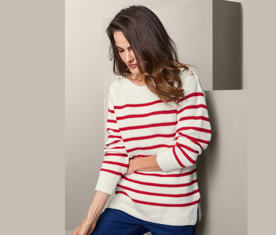 Žena pózuje v pleteném svetru s knoflíky s červenými pruhy a džínách.