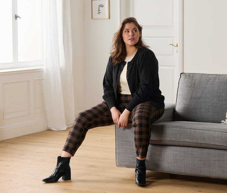 Žena sedí na šedé pohovce a má na sobě krémové tričko, kostkované strečové kalhoty a kabátek z jemného úpletu s objemnými rukávy.