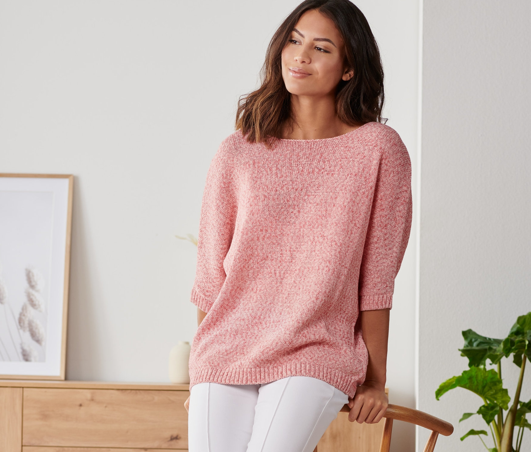 Žena opřená o židli má na sobě letní pletený svetr z páskové příze, bílé kalhoty a tenisky.