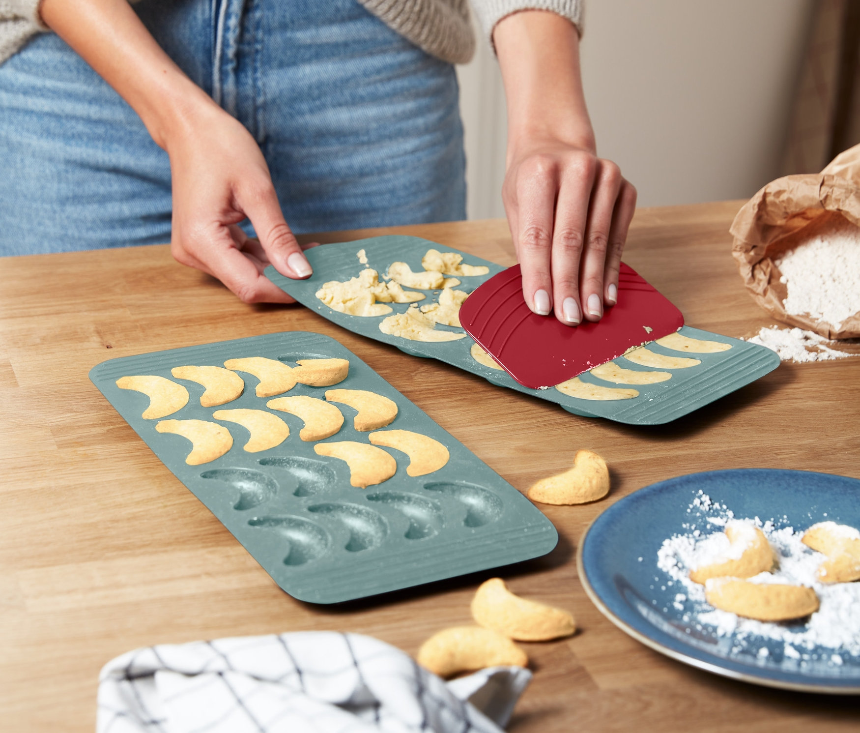 Žena pomocí červené špachtle odděluje těsto z dvou zelených forem na pečení vanilkových rohlíčků.