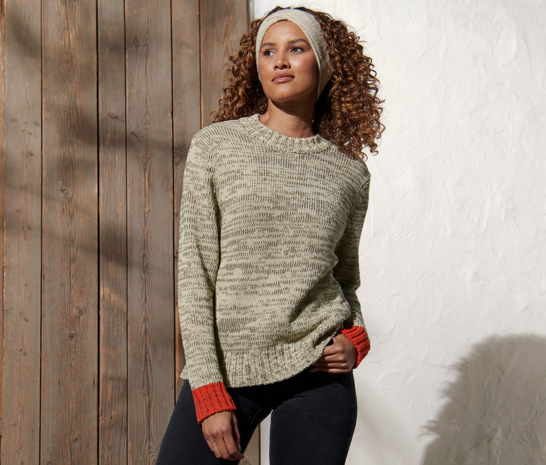 Žena s kudrnatými vlasy pózuje v svetru z hrubé pleteniny a džínových kalhotách.
