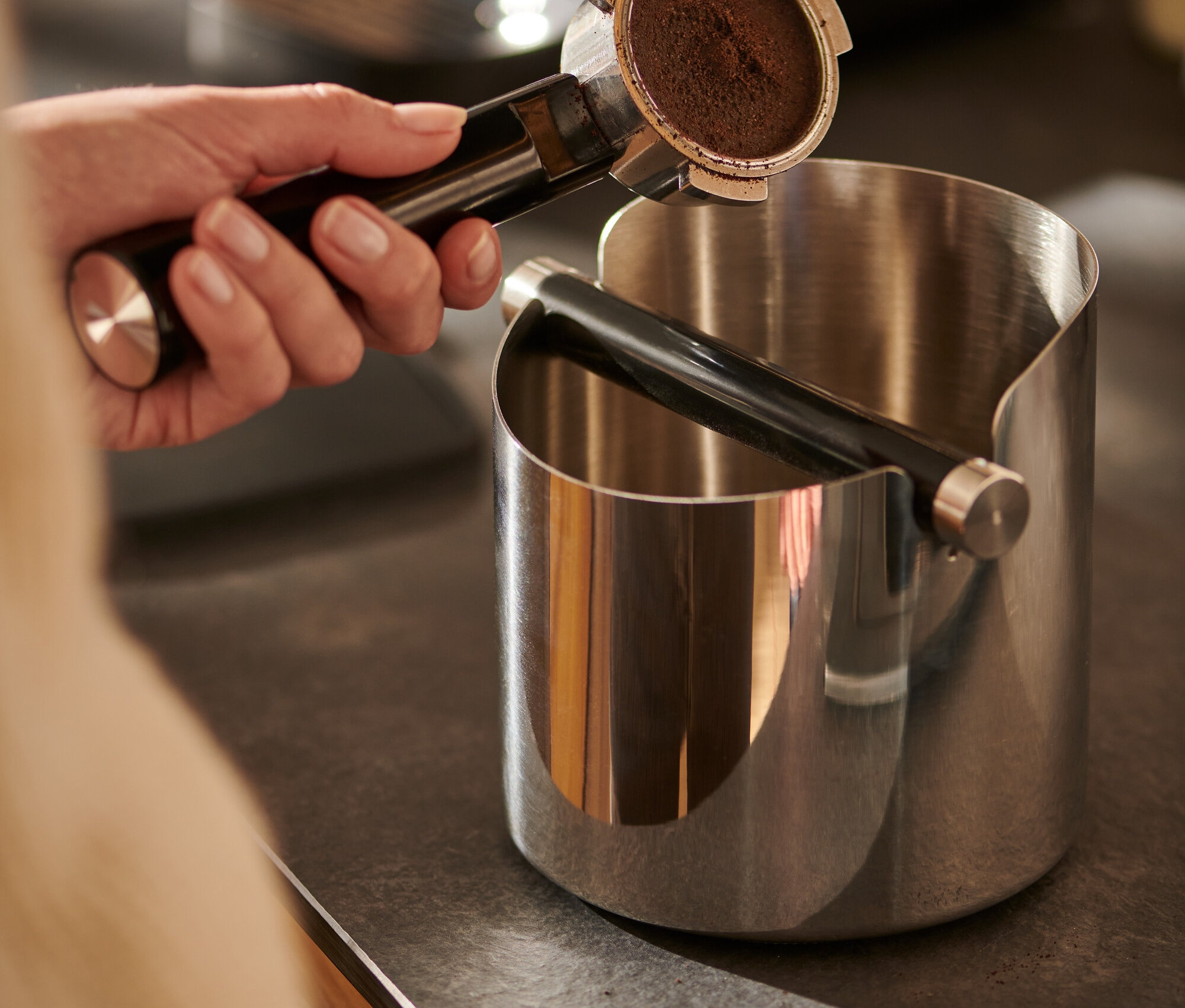 Ruka drží páku s kávou nad lesklým vyklepávacím boxem.