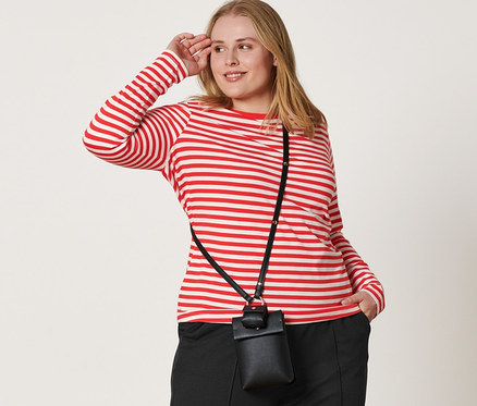 Žena pózuje v červeno-bílém pruhovaném tričku s dlouhým rukávem, černých teplákových kalhotách s puky, kožených teniskách a s mini taštičkou přes rameno.